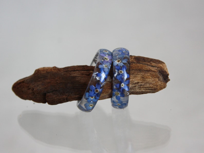 May include: Two clear resin bangles with blue forget-me-not flowers and silver flakes embedded inside. The bangles are displayed on a piece of driftwood against a white background. The floral design adds a touch of nature-inspired elegance.