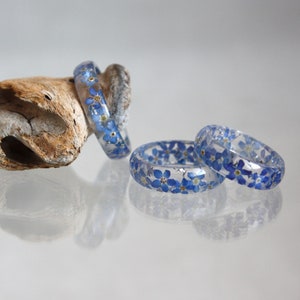 May include: Clear resin rings with embedded blue forget-me-not flowers. The rings are displayed on a light-colored surface, with one resting on a piece of driftwood. The rings are approximately 0.75 inches wide.