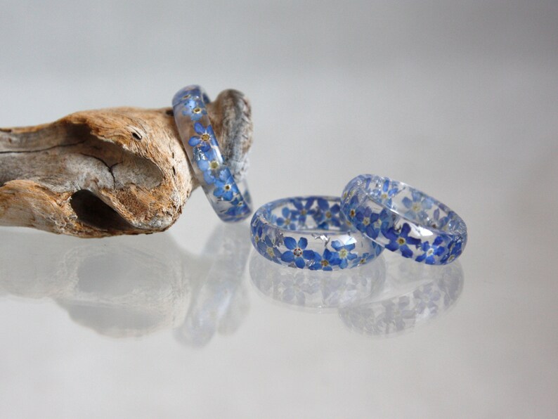May include: Clear resin rings with embedded blue forget-me-not flowers. The rings are displayed on a light-colored surface, with one resting on a piece of driftwood. The rings are approximately 0.75 inches wide.