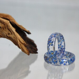 May include: Two clear resin rings embedded with small blue flowers. The rings are stacked on top of each other, reflecting light. A piece of driftwood is in the background, adding a natural element to the composition.