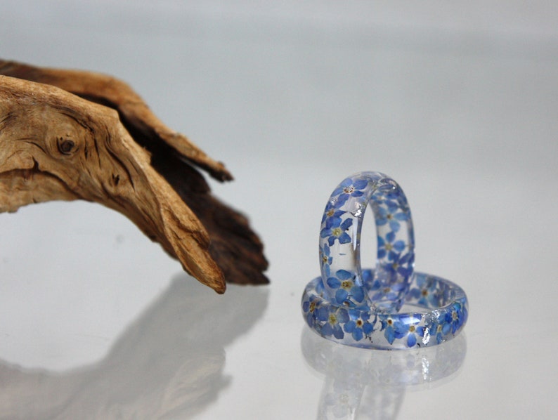 May include: Two clear resin rings embedded with small blue flowers. The rings are stacked on top of each other, reflecting light. A piece of driftwood is in the background, adding a natural element to the composition.