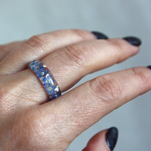 May include: A clear resin ring with small blue flowers and silver flecks embedded within. The ring is worn on a finger, showcasing the delicate floral design. The ring's band is approximately 0.25 inches wide.