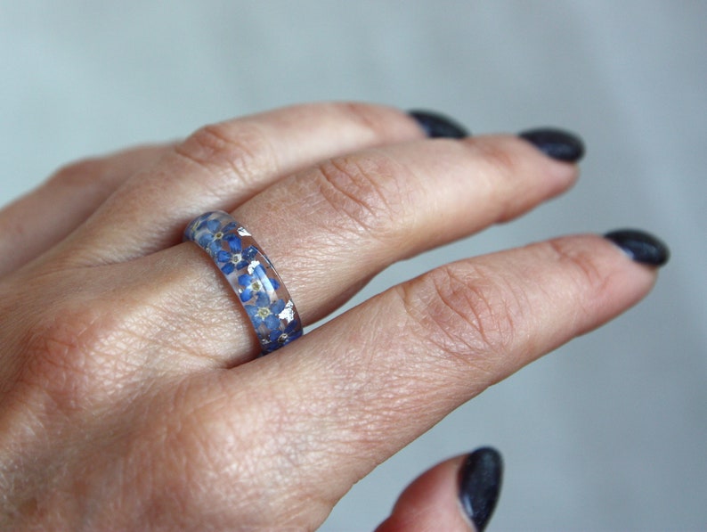 May include: A clear resin ring with small blue flowers and silver flecks embedded within. The ring is worn on a finger, showcasing the delicate floral design. The ring's band is approximately 0.25 inches wide.