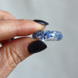 May include: A clear resin ring embedded with small blue flowers. The ring is held between fingers, showcasing the delicate floral design. The ring's band is approximately 0.25 inches wide, with the flowers visible throughout the clear resin.