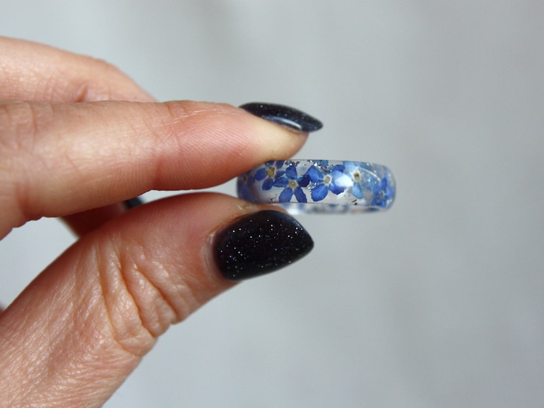 May include: A clear resin ring embedded with small blue flowers. The ring is held between fingers, showcasing the delicate floral design. The ring's band is approximately 0.25 inches wide, with the flowers visible throughout the clear resin.