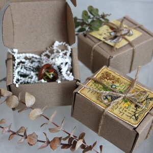 May include: Three small, brown cardboard boxes with twine and paper tags. The boxes are filled with small, handmade glass ornaments. The boxes are sitting on a white surface.