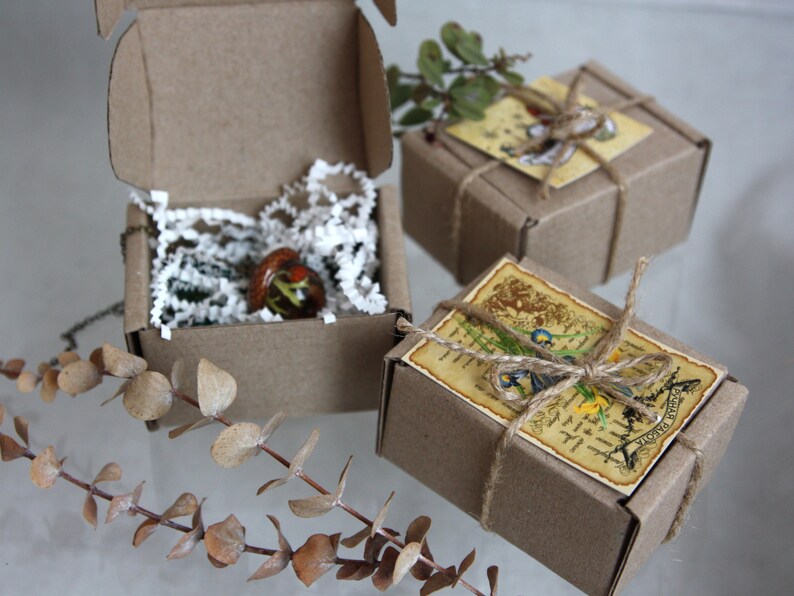May include: Three small, brown cardboard boxes with twine and paper tags. The boxes are filled with small, handmade glass ornaments. The boxes are sitting on a white surface.