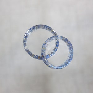 May include: Two clear resin rings, each embedded with small blue and yellow floral elements. The rings are interlocked, creating a unique design. The transparent material allows the floral details to be fully visible, offering a delicate and natural aesthetic.