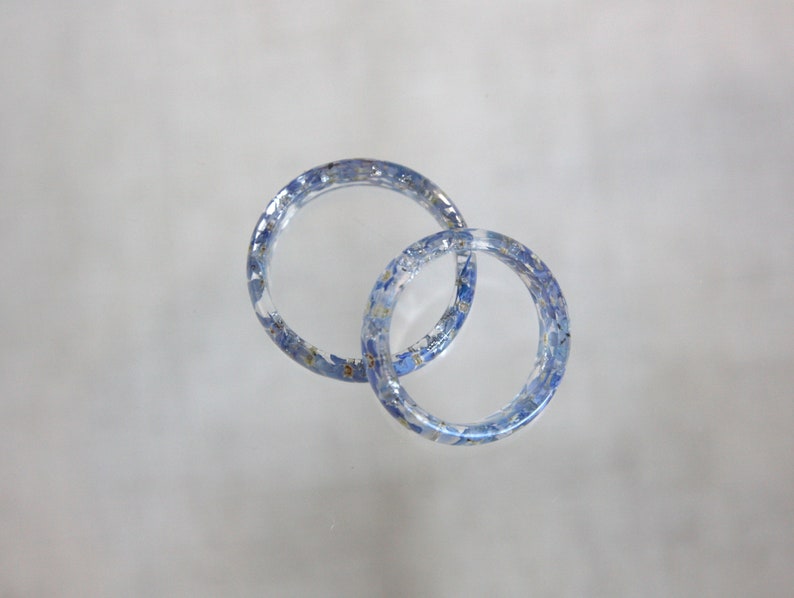 May include: Two clear resin rings, each embedded with small blue and yellow floral elements. The rings are interlocked, creating a unique design. The transparent material allows the floral details to be fully visible, offering a delicate and natural aesthetic.