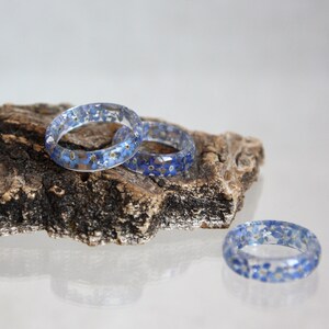 May include: Three clear resin rings with embedded blue and yellow flowers rest on a piece of wood. The rings are circular and showcase the delicate floral inclusions, creating a unique and natural aesthetic. The rings are a beautiful accessory.