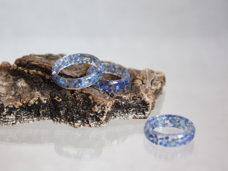 May include: Three clear resin rings with embedded blue and yellow flowers rest on a piece of wood. The rings are circular and showcase the delicate floral inclusions, creating a unique and natural aesthetic. The rings are a beautiful accessory.