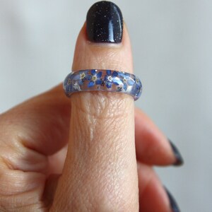 May include: A clear resin ring with blue forget-me-not flowers and silver flakes embedded within. The ring is worn on a finger, showcasing the floral design. The ring's band is approximately 0.25 inches wide.