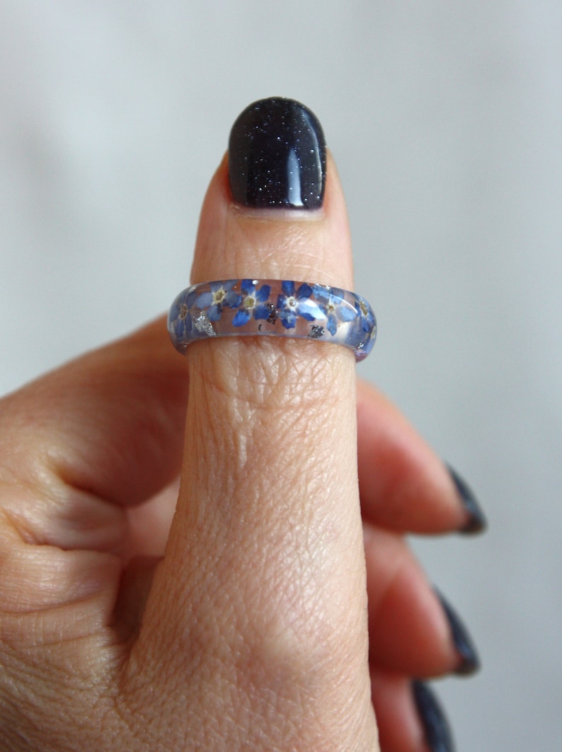 May include: A clear resin ring with blue forget-me-not flowers and silver flakes embedded within. The ring is worn on a finger, showcasing the floral design. The ring's band is approximately 0.25 inches wide.