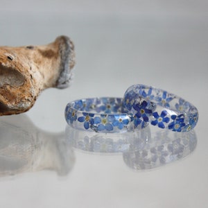 May include: Two clear resin rings with embedded blue forget-me-not flowers. The rings are displayed on a reflective surface, showcasing the delicate floral design. A piece of driftwood is in the background.