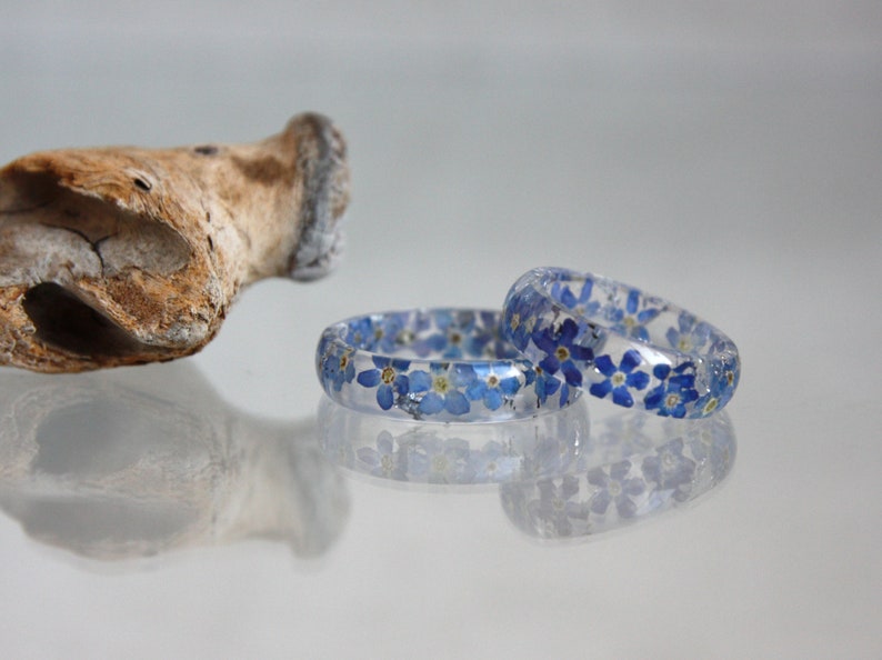 May include: Two clear resin rings with embedded blue forget-me-not flowers. The rings are displayed on a reflective surface, showcasing the delicate floral design. A piece of driftwood is in the background.