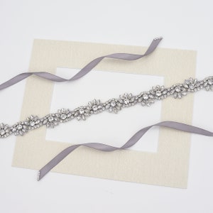 May include: A decorative belt featuring a continuous row of clear, faceted crystals arranged in floral-inspired clusters. The crystal embellishment is attached to a smooth, grey satin ribbon that extends from both ends of the jeweled section.