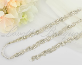 May include: A silver bridal sash featuring a beaded and rhinestone design. The sash is adorned with floral motifs and has a delicate, sparkling finish.