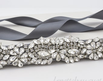 May include: A grey satin ribbon sash with a crystal embellishment. The embellishment is made of clear rhinestones in a floral design.