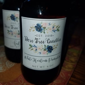 May include: Two brown glass bottles with white labels. The label on the front bottle reads "EST. 2018. Dove Tree Candles White Heirloom Pumpkin NET WT. 4 OZ". The label features a floral design with blue and pink flowers.
