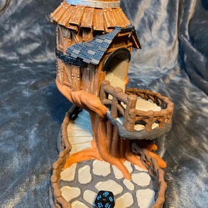 Puede incluir: Una torre de dados de madera en forma de casa en el árbol con un esquema de color marrón, gris y negro. La torre tiene un pequeño techo y un balcón. La base de la torre es un tronco de árbol con una escalera de caracol que conduce a la torre. La base tiene una superficie de piedra con un dado negro sobre ella.
