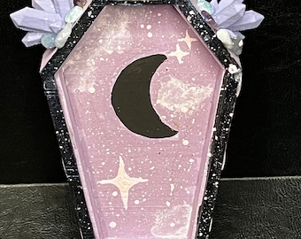 May include: A small purple coffin-shaped trinket box with a crescent moon and stars painted on the lid. The box is decorated with purple and blue crystals.