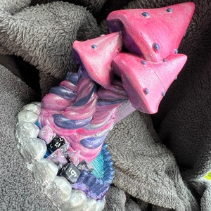 May include: A purple and pink mushroom-shaped dice tower with a swirling design. The tower has a base with a silver and blue design and includes multiple black and purple dice.