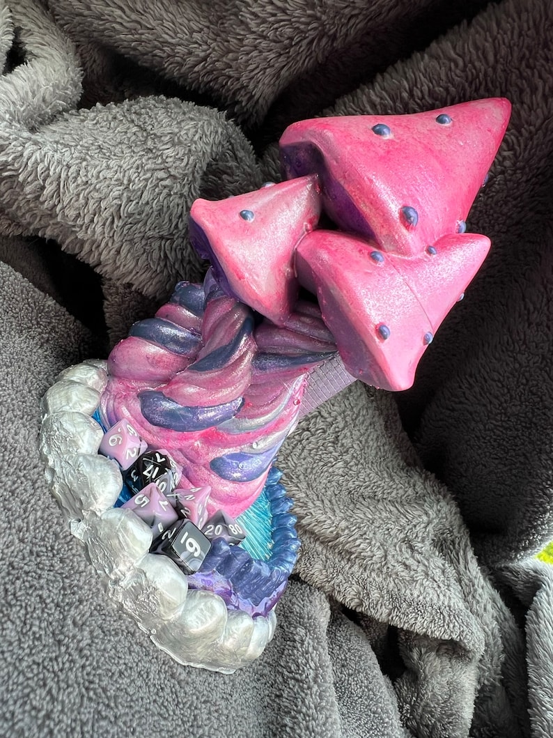 May include: A purple and pink mushroom-shaped dice tower with a swirling design. The tower has a base with a silver and blue design and includes multiple black and purple dice.