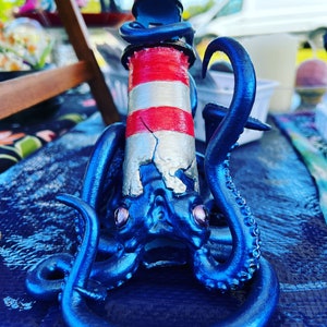 Könnte beinhalten: Eine blaue Oktopus-Figur mit einem rot-weiß gestreiften Leuchtturm auf dem Rücken. Der Oktopus hat große, runde Augen und ist aus glänzendem, metallischem Material gefertigt.