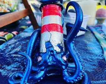 May include: A blue octopus figurine with a red and white striped lighthouse on its back. The octopus has large, round eyes and is made of a shiny, metallic material.