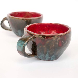 May include: Two handmade ceramic mugs with a dark brown glaze and red interior. The mugs have a unique, speckled design.