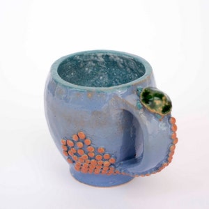 May include: A light blue ceramic mug with a textured handle and a green and white glaze detail. The mug has a speckled design with orange dots.