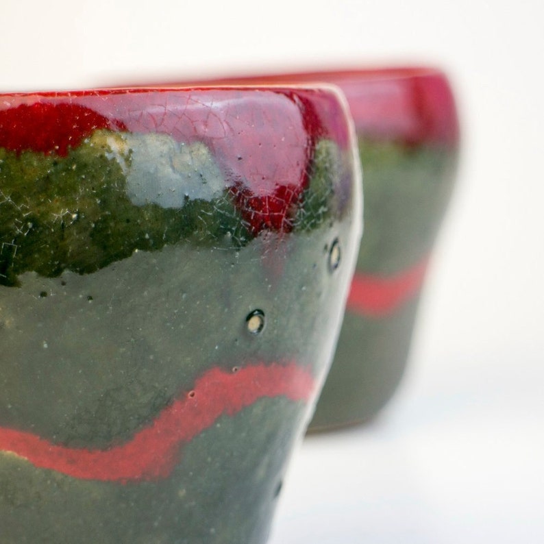May include: Two ceramic mugs with a green and red glaze. The mugs have a crackled finish and a unique, abstract design.