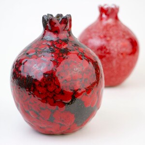 May include: Two red ceramic pomegranate ornaments with black detailing. The ornament on the left has a more textured surface.
