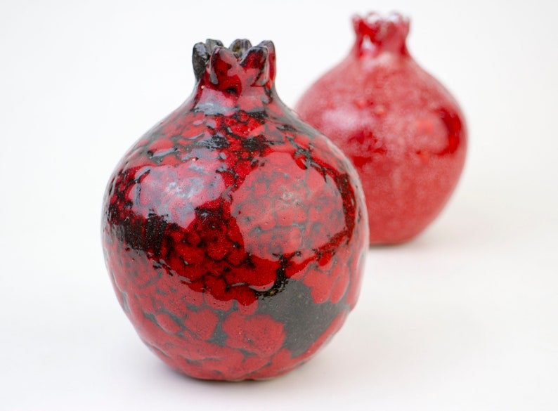 May include: Two red ceramic pomegranate ornaments with black detailing. The ornament on the left has a more textured surface.