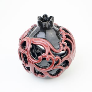 Puede incluir: Una granada decorativa de cerámica con un acabado negro brillante y adornos rojos. El objeto en forma de fruta tiene una parte superior en forma de corona y un diseño calado, creando una estética única. Una llamativa pieza de decoración del hogar.