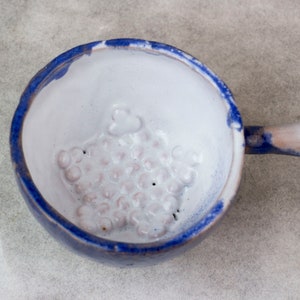 Puede incluir: Un tazón de cerámica blanca con un borde azul y un asa. El tazón tiene una superficie texturizada con pequeños agujeros.