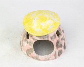 May include: A ceramic house shaped like a mushroom with a pink and grey base and a yellow top. The house has several holes in the base.
