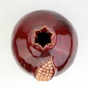 May include: A ceramic pomegranate vase with a glossy, deep red glaze. The top opening is star-shaped with gold trim, and a section of the fruit's skin is detailed with a textured, tan pattern. The vase is a decorative piece.