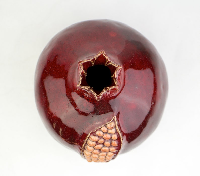 May include: A ceramic pomegranate vase with a glossy, deep red glaze. The top opening is star-shaped with gold trim, and a section of the fruit's skin is detailed with a textured, tan pattern. The vase is a decorative piece.