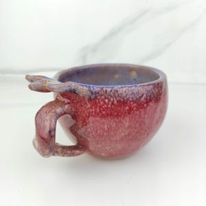 Puede incluir: Una taza de cerámica hecha a mano con un esmalte rojo y morado texturizado único. La taza tiene un asa con forma de cabeza de dragón.