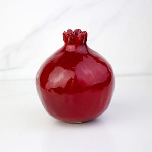 May include: A glossy, deep red ceramic pomegranate sculpture. The fruit-shaped object has a textured crown and a smooth, reflective surface. The pomegranate is a decorative item, likely for home decor.
