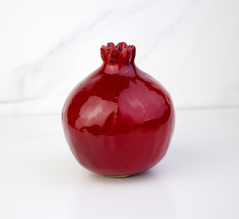 May include: A glossy, deep red ceramic pomegranate sculpture. The fruit-shaped object has a textured crown and a smooth, reflective surface. The pomegranate is a decorative item, likely for home decor.