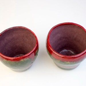 May include: Two small ceramic cups with red, green and brown glaze. The cups are empty and sitting on a white surface.