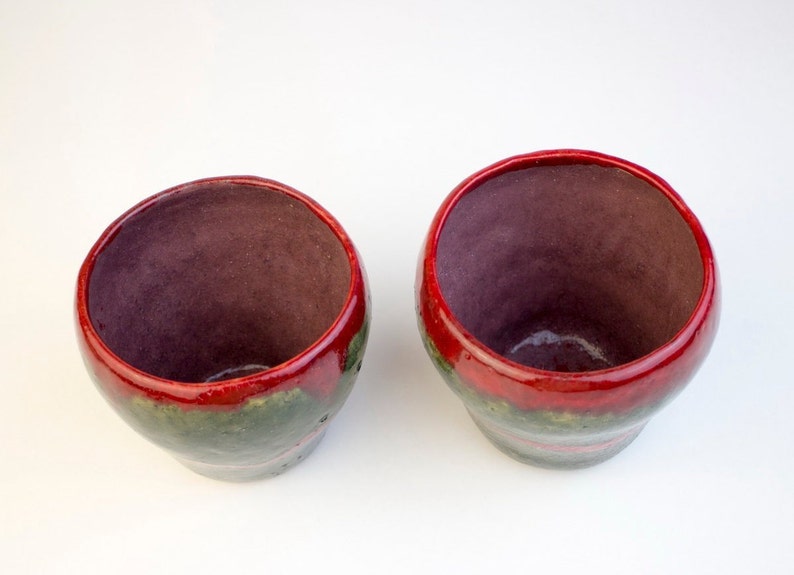 May include: Two small ceramic cups with red, green and brown glaze. The cups are empty and sitting on a white surface.