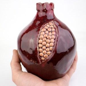 May include: A ceramic pomegranate sculpture in a deep burgundy glaze. The fruit is detailed with a carved section revealing the seeds, and a textured surface. The pomegranate is held in a hand against a white background.