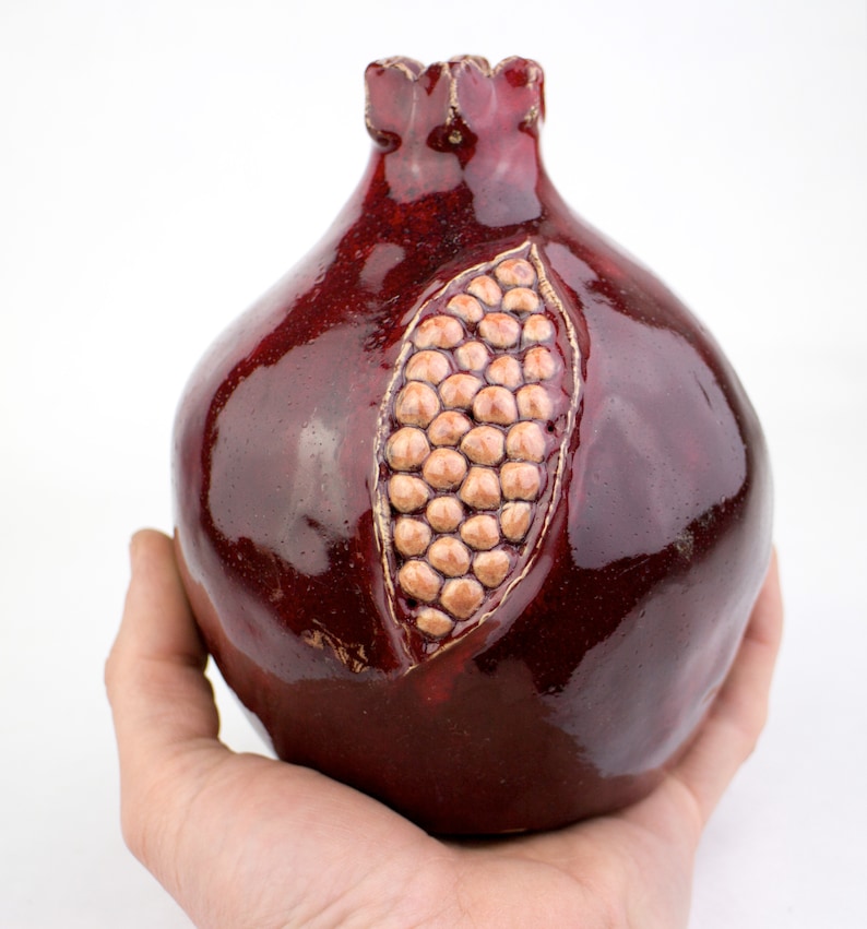 May include: A ceramic pomegranate sculpture in a deep burgundy glaze. The fruit is detailed with a carved section revealing the seeds, and a textured surface. The pomegranate is held in a hand against a white background.
