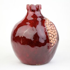 May include: A ceramic pomegranate-shaped vase with a glossy, deep red glaze. The vase has a section carved out to reveal a cluster of tan-colored seeds. The top of the vase has a textured, ruffled edge.