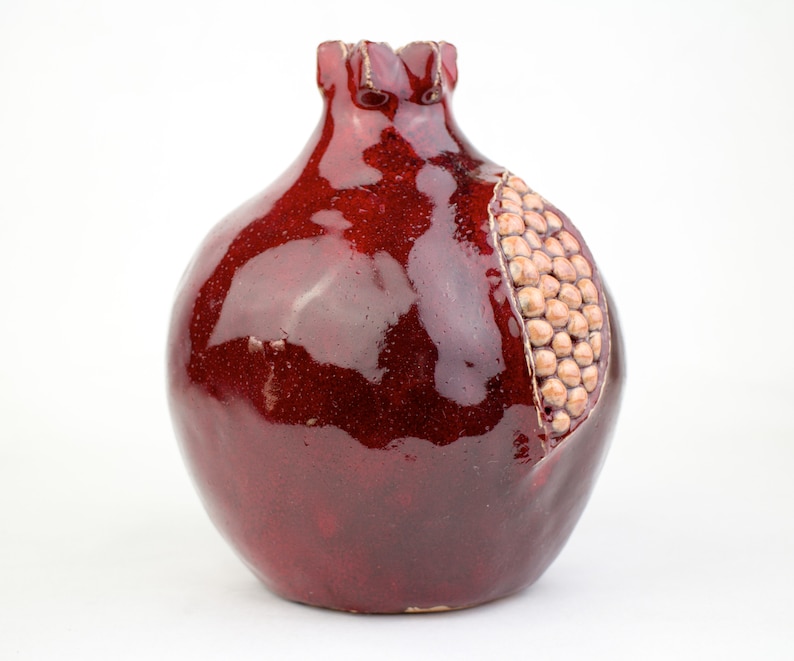 May include: A ceramic pomegranate-shaped vase with a glossy, deep red glaze. The vase has a section carved out to reveal a cluster of tan-colored seeds. The top of the vase has a textured, ruffled edge.
