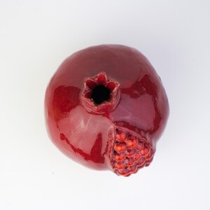 May include: A glossy, ceramic pomegranate in a deep red hue. The fruit features a detailed, textured seed section and a star-shaped calyx. The pomegranate is a decorative item, likely for home decor or as a unique kitchen accent.