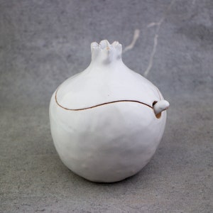 May include: White ceramic sugar bowl shaped like a pomegranate with a lid and a small spout. The bowl has a subtle glaze and a slightly textured surface.